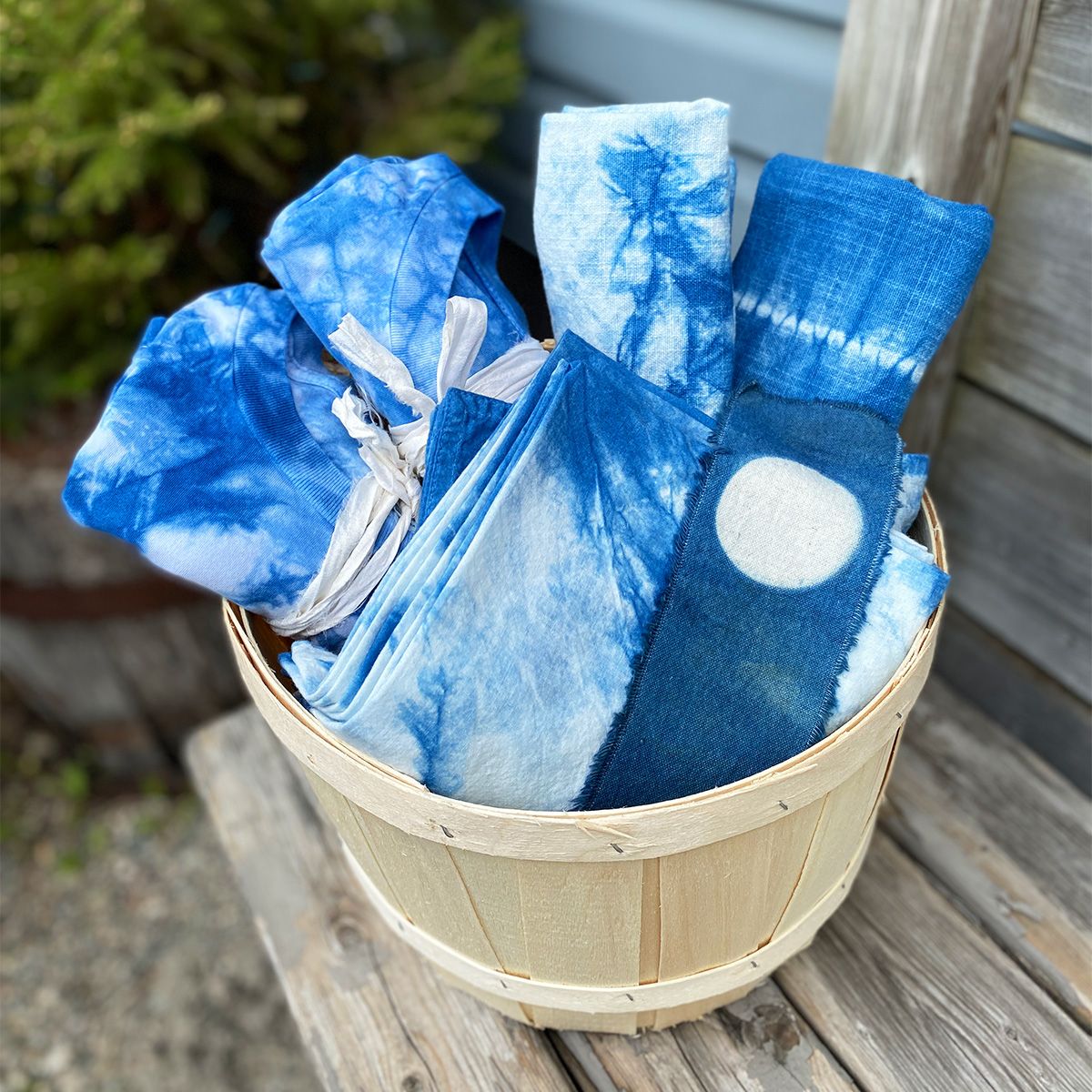 A basket on a table outdoors filled with folded indigo-dyed T-shirts, bookmarks and other textile products