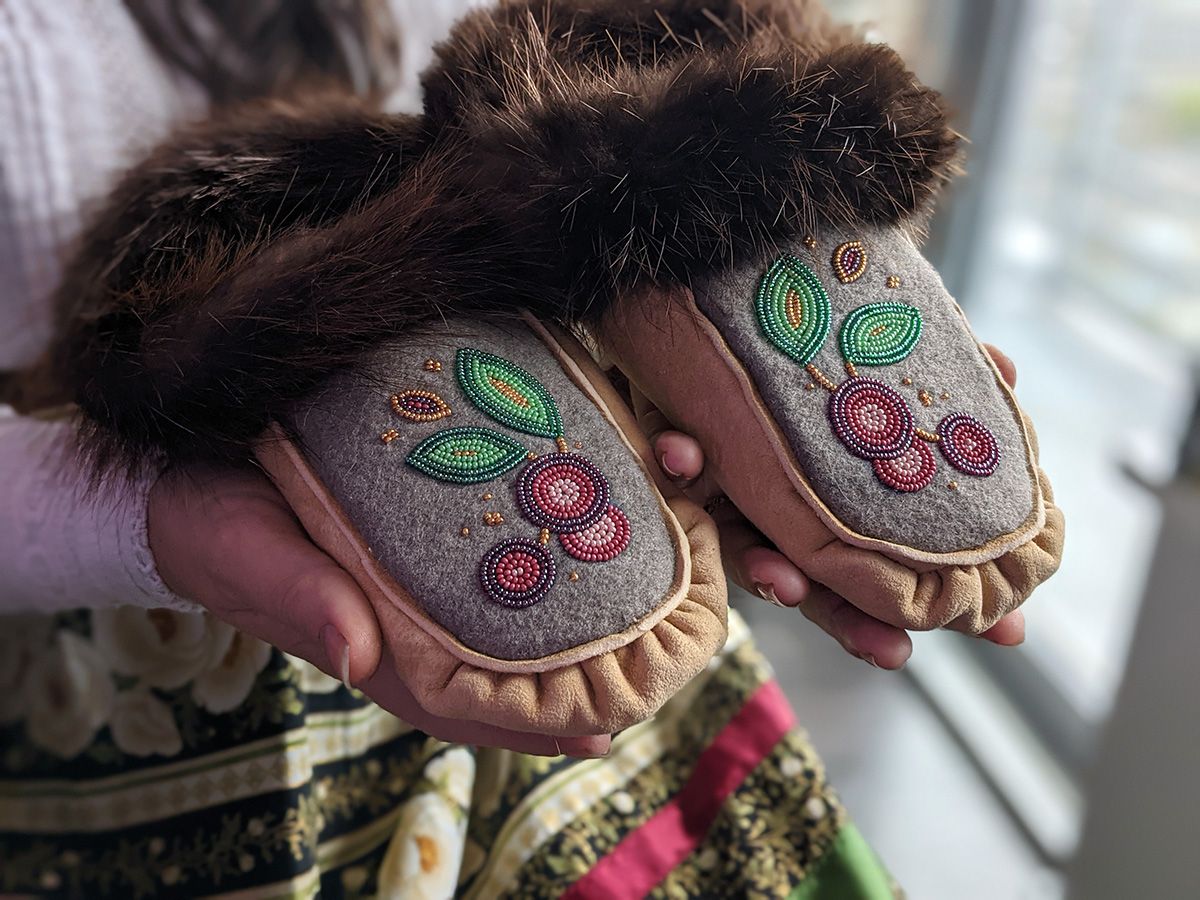 A person wearing a ribbon skirt holding up a pair of Indigenous Nouveau beaded moccasins with fur trim.