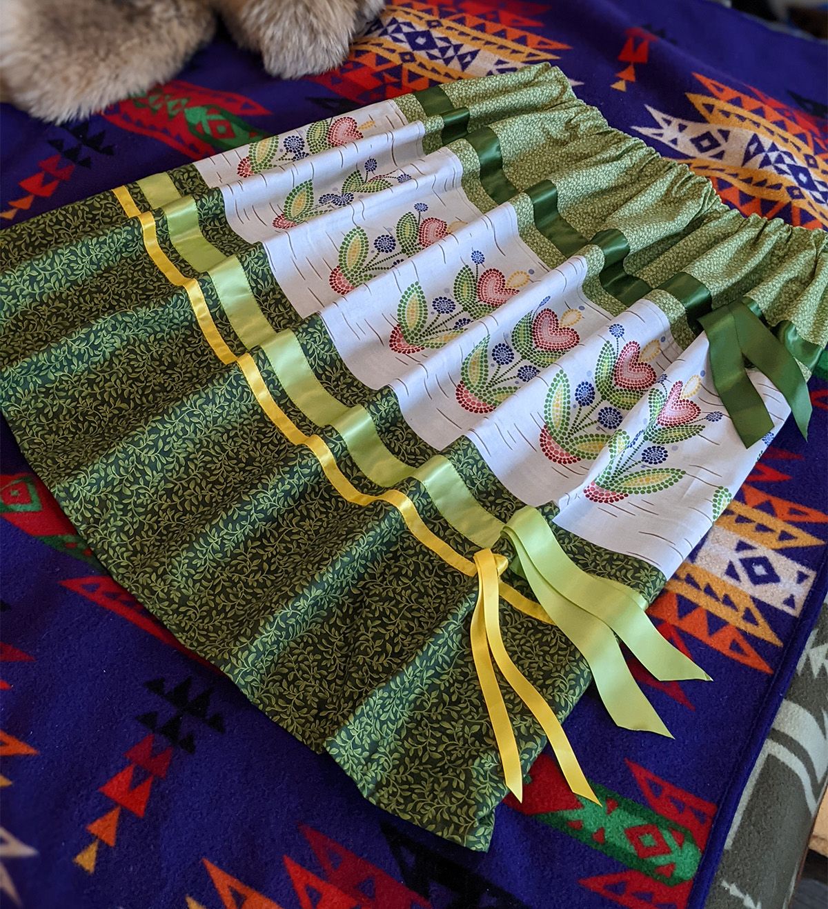 A green, yellow and white ribbon skirt with Indigenous Nouveau fabric on a blanket-covered table