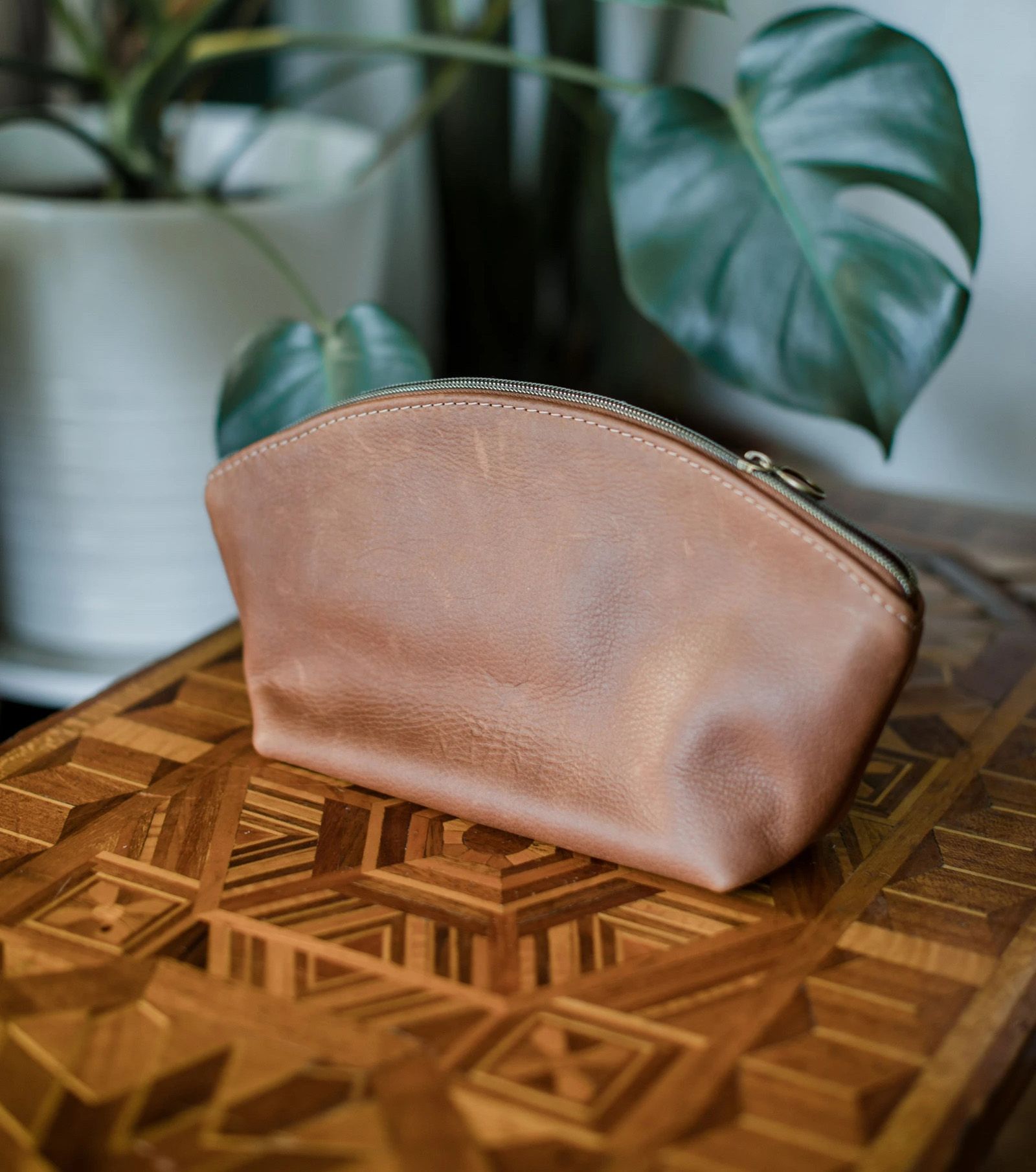 A brown leather cosmetics bag with a zipper on a wooden table with inlaid design, with a plant in the background.