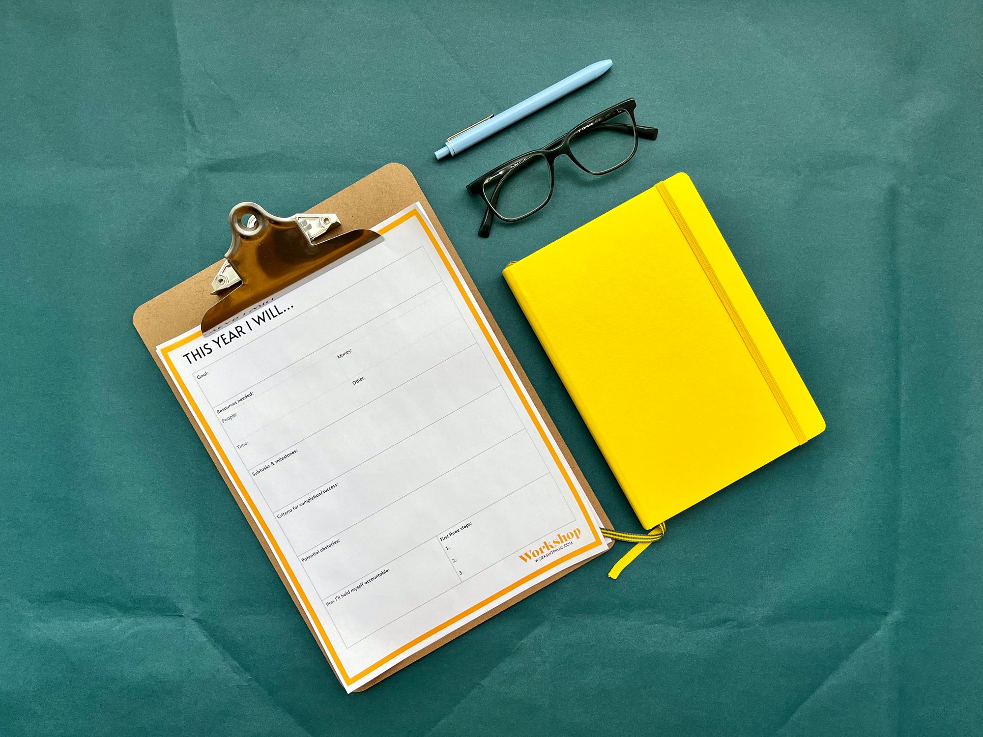 A notebook, a pair of glasses, a pen, and a clipboard holding a goal-setting workshee