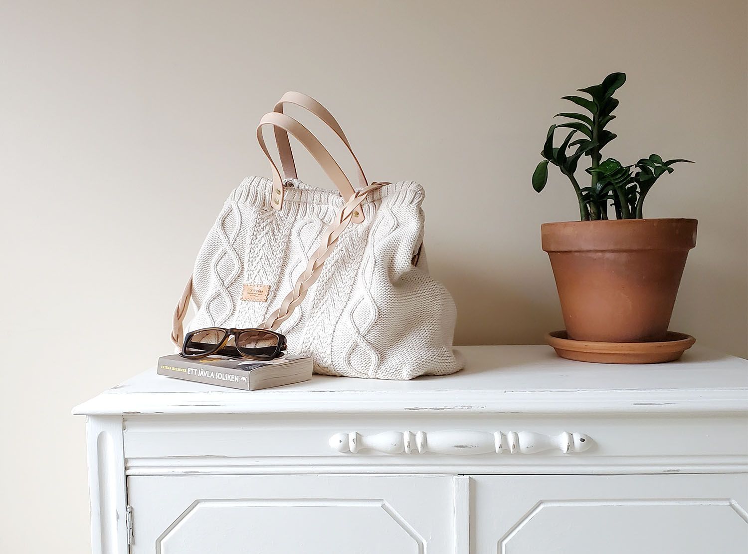 A cream-coloured cable-knit bag on a white dresser with sunglasses, a book and a potted plant