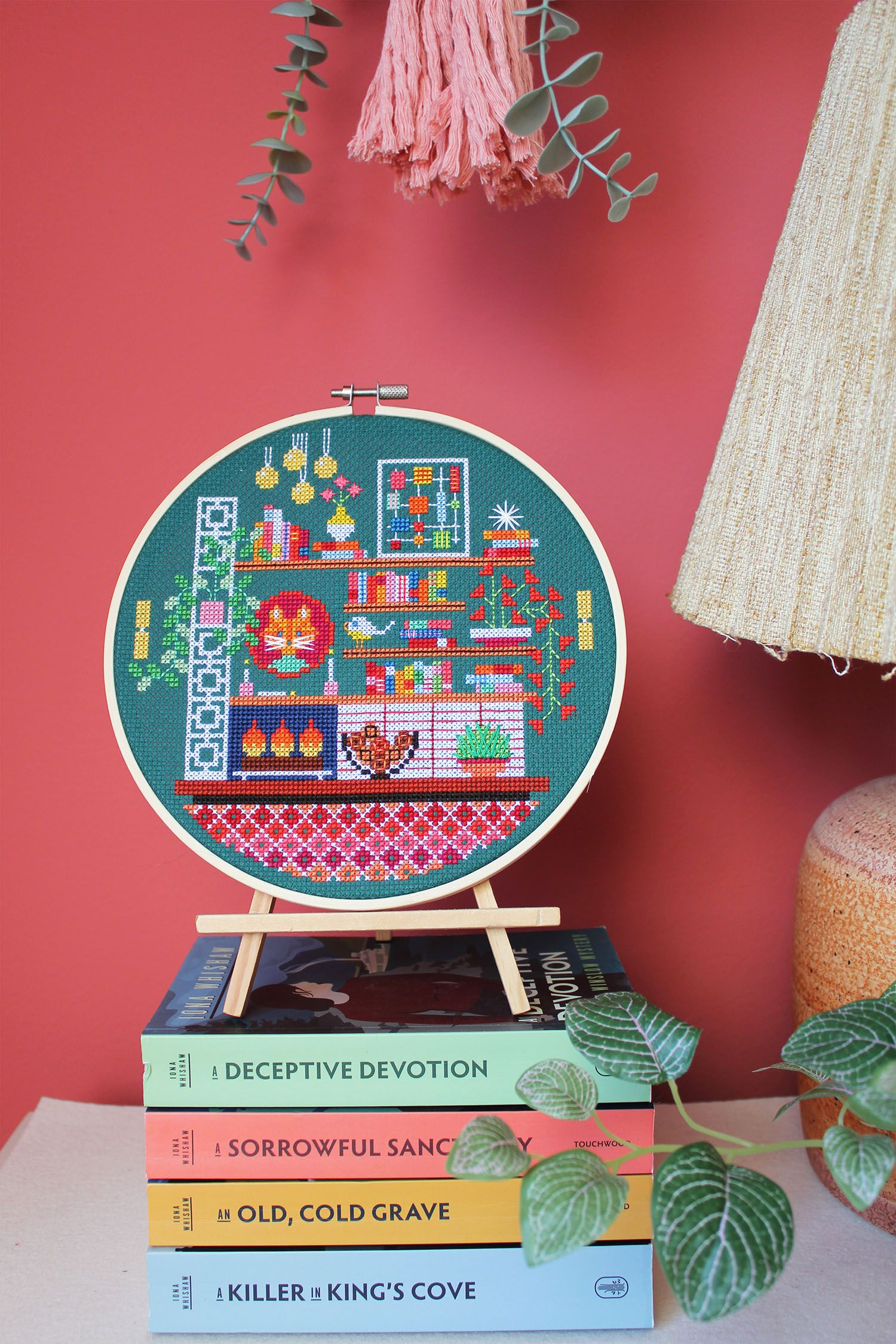 A framed cross-stitch on a stand on top of a pile of books, on a dresser with a plant and lamp