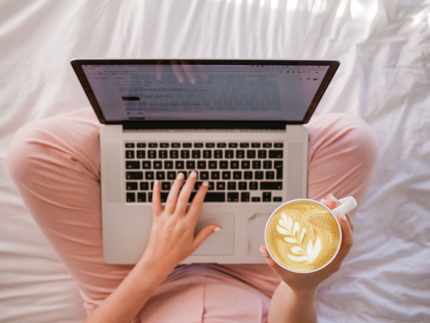 A top-down view of someone in a pink onesie sitting cross-legged on white sheets with a laptop in their lap, holding a cappuccino