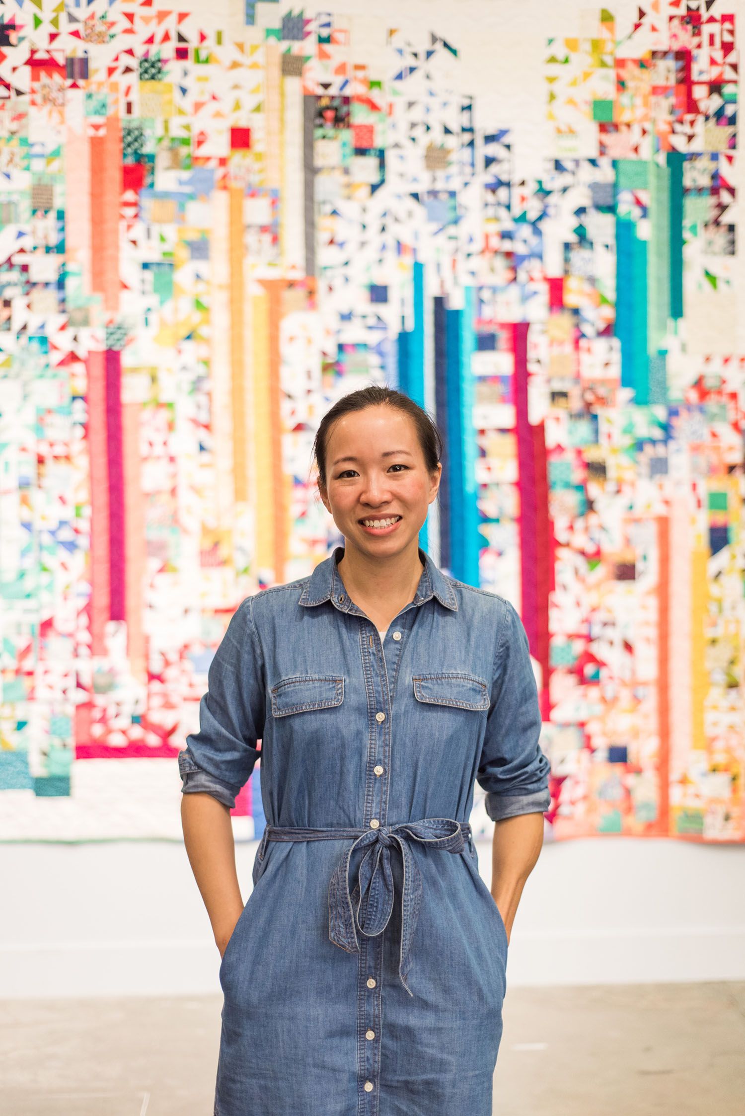 A woman in a denim dress standing in a gallery with a quilt in the background