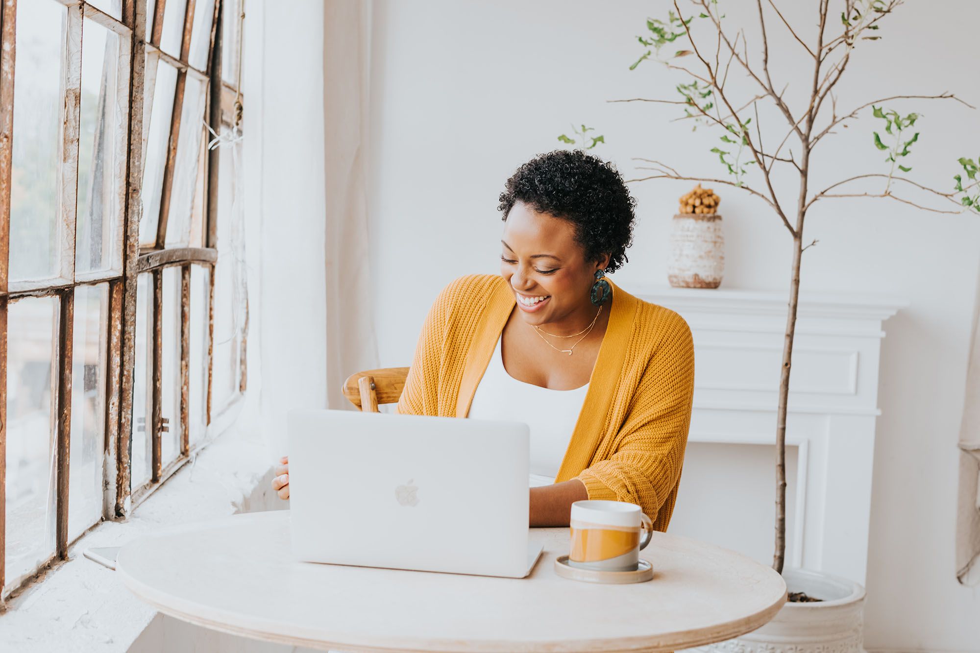 Jasmine Williams wearing a white top and yellow cardigan, sitting at a laptop next to a window