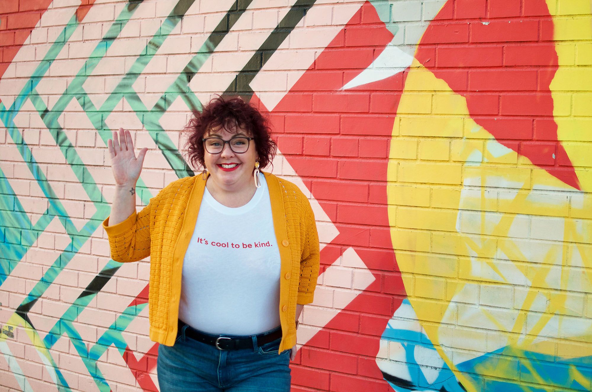 Kim Cota standing in front of a colourful mural wall, smiling and waving one hand