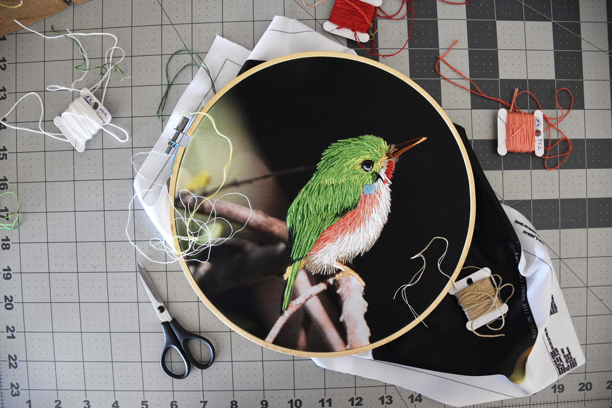 A work table with embroidery supplies and a project in a hoop, of a bird