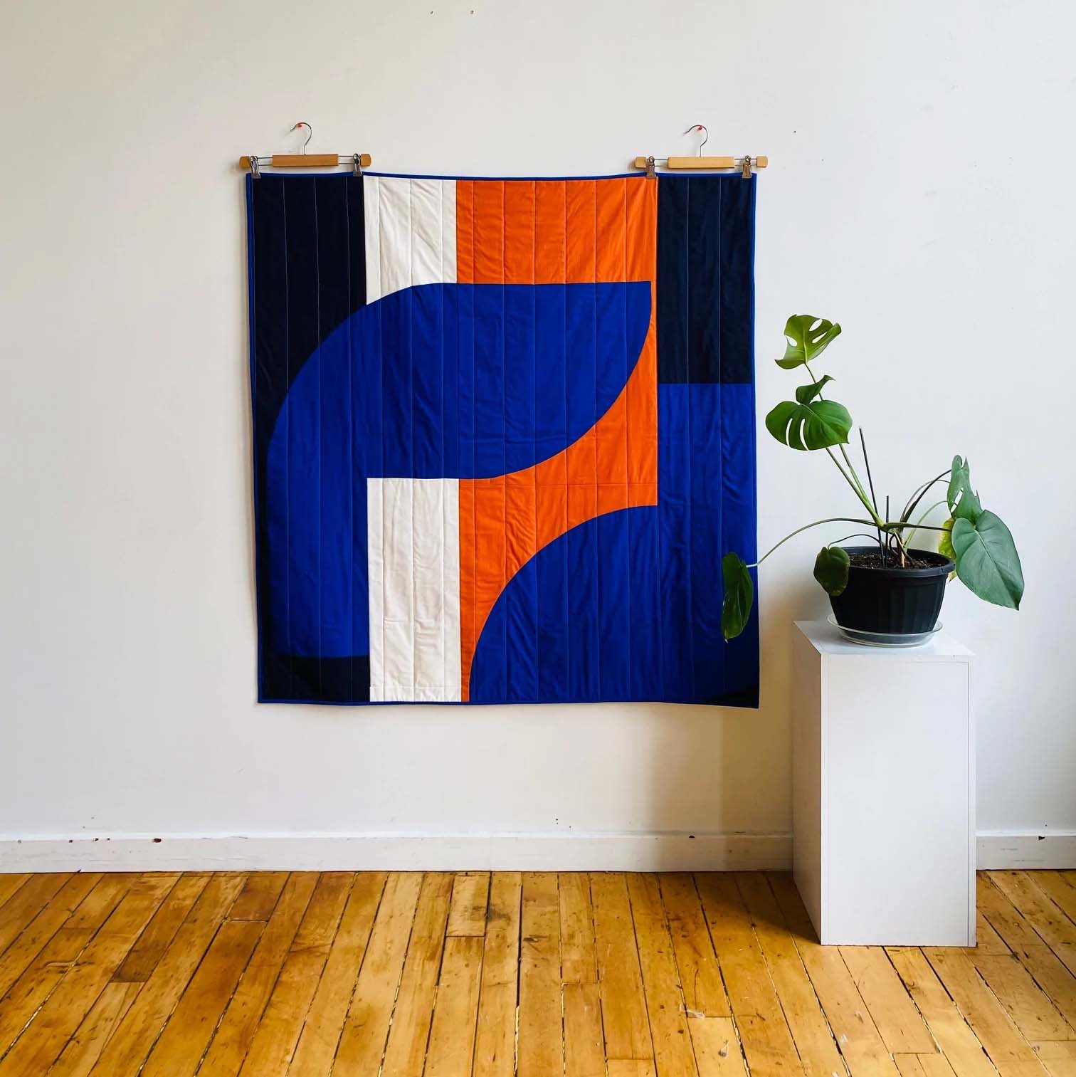 Abstract quilt in blue, orange, black and white hanging on white wall