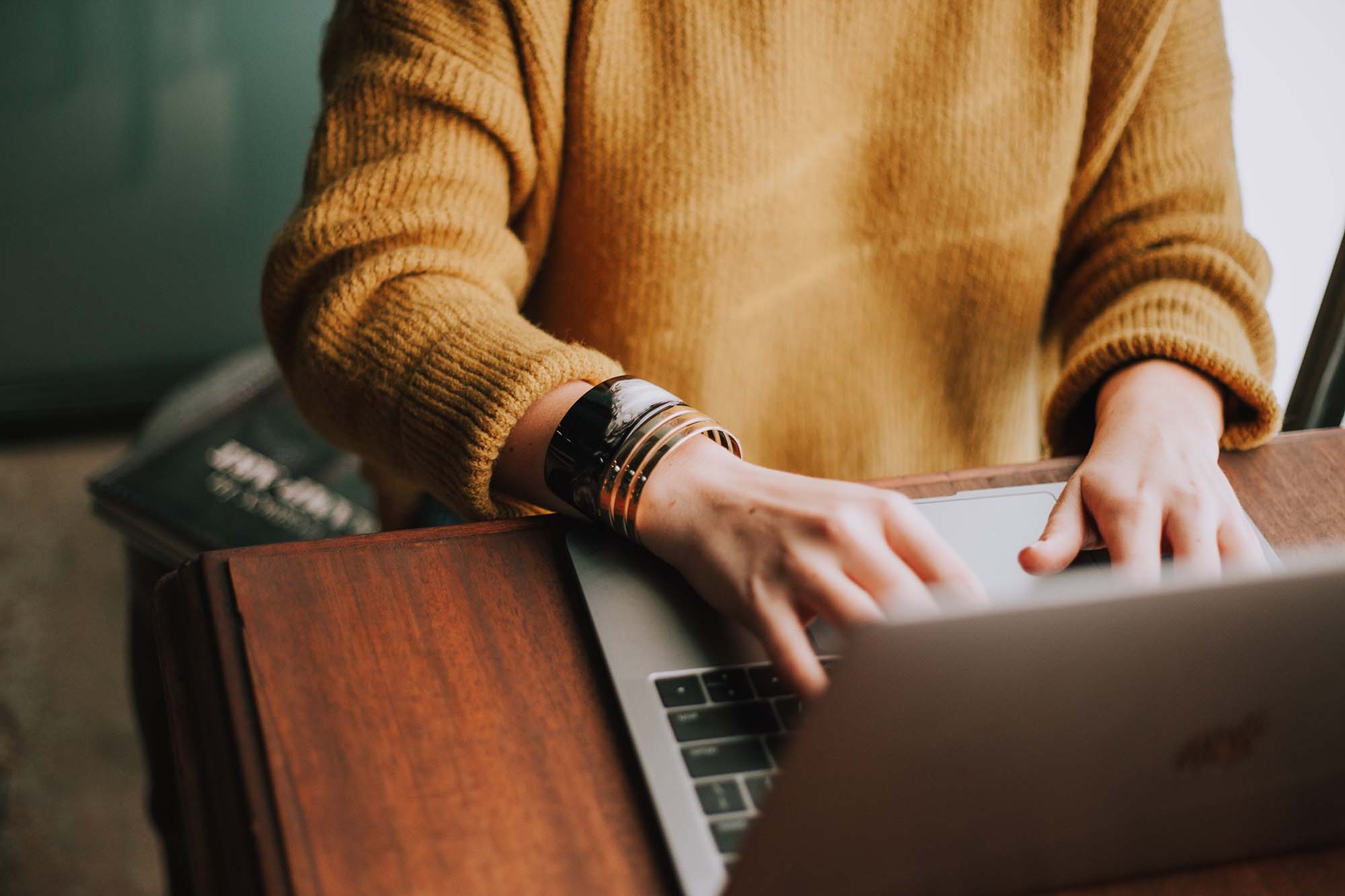 Person in mustard-coloured sweater sitting at a wooden table typing on a laptop computer