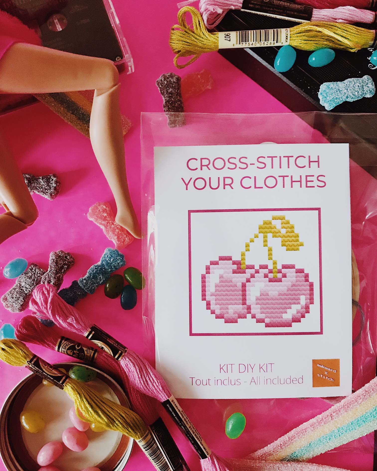 cross-stitch kit of cherries with embroidery floss on a hot-pink background surrounded by candy