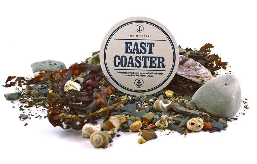 A cardboard coaster printed with text surrounded by rocks, shells and seaweed