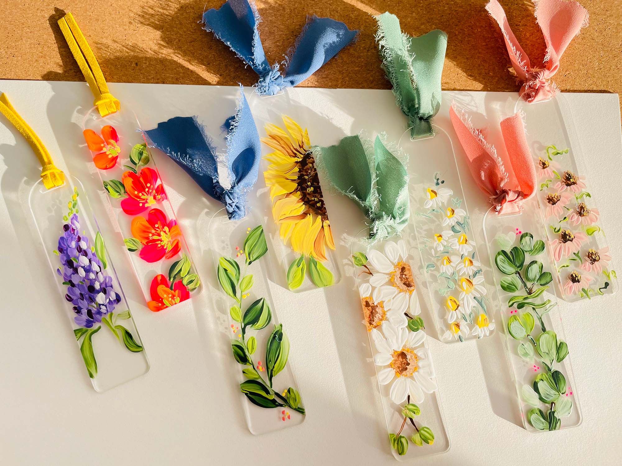 A row of flower-painted acrylic bookmarks on a table in sunlight