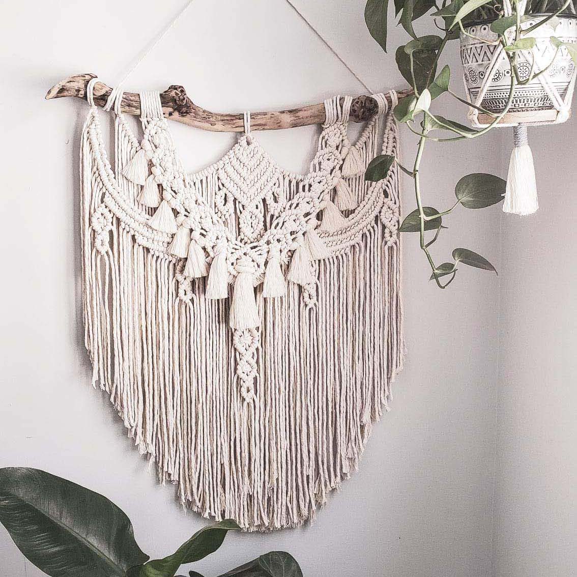 White macrame piece hanging on a wall with plants surrounding it