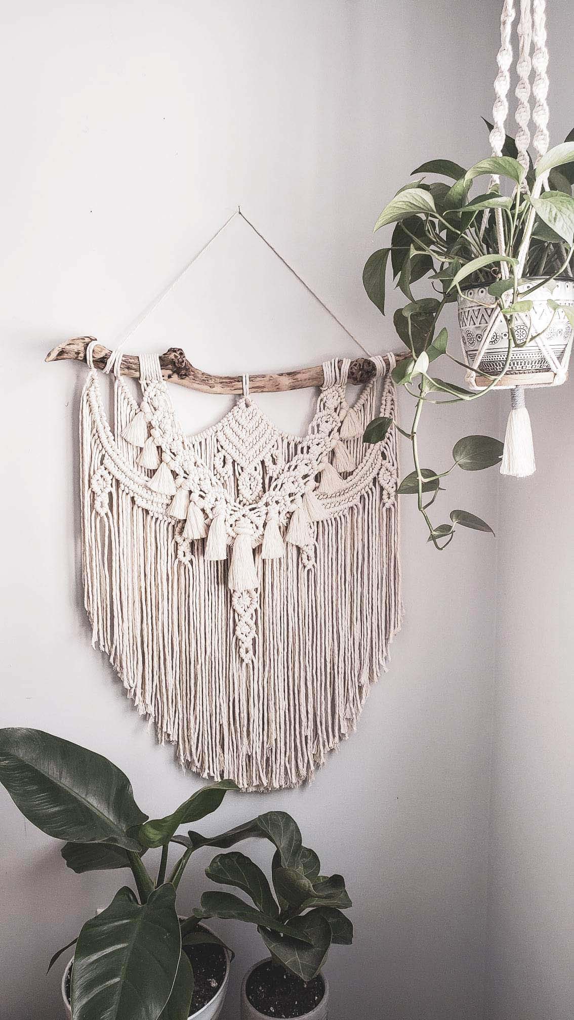 A white macrame wall hanging on a wall surrounded by plants