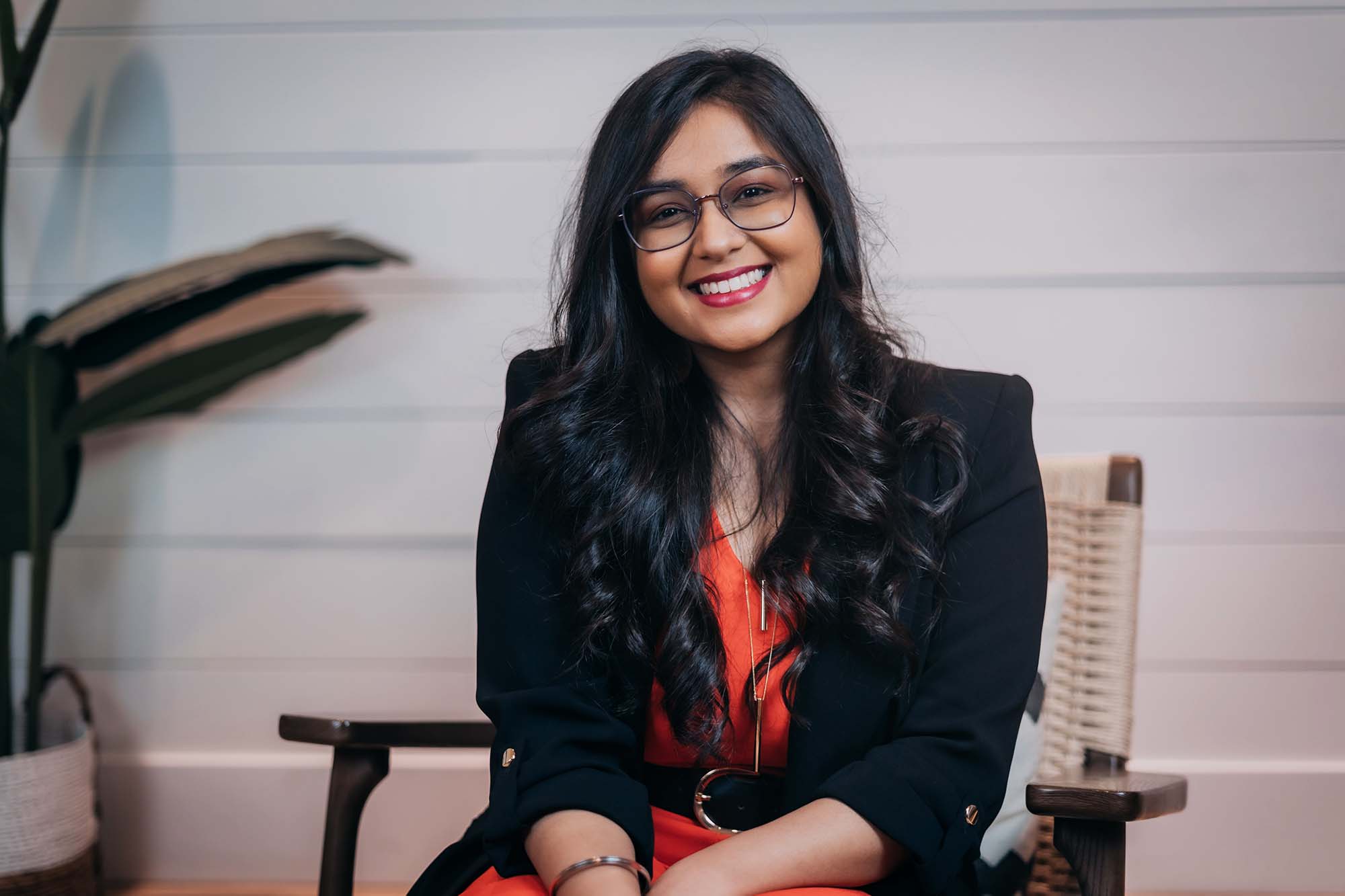 A person in business attire and glasses sits in a chair and smiles at the camera